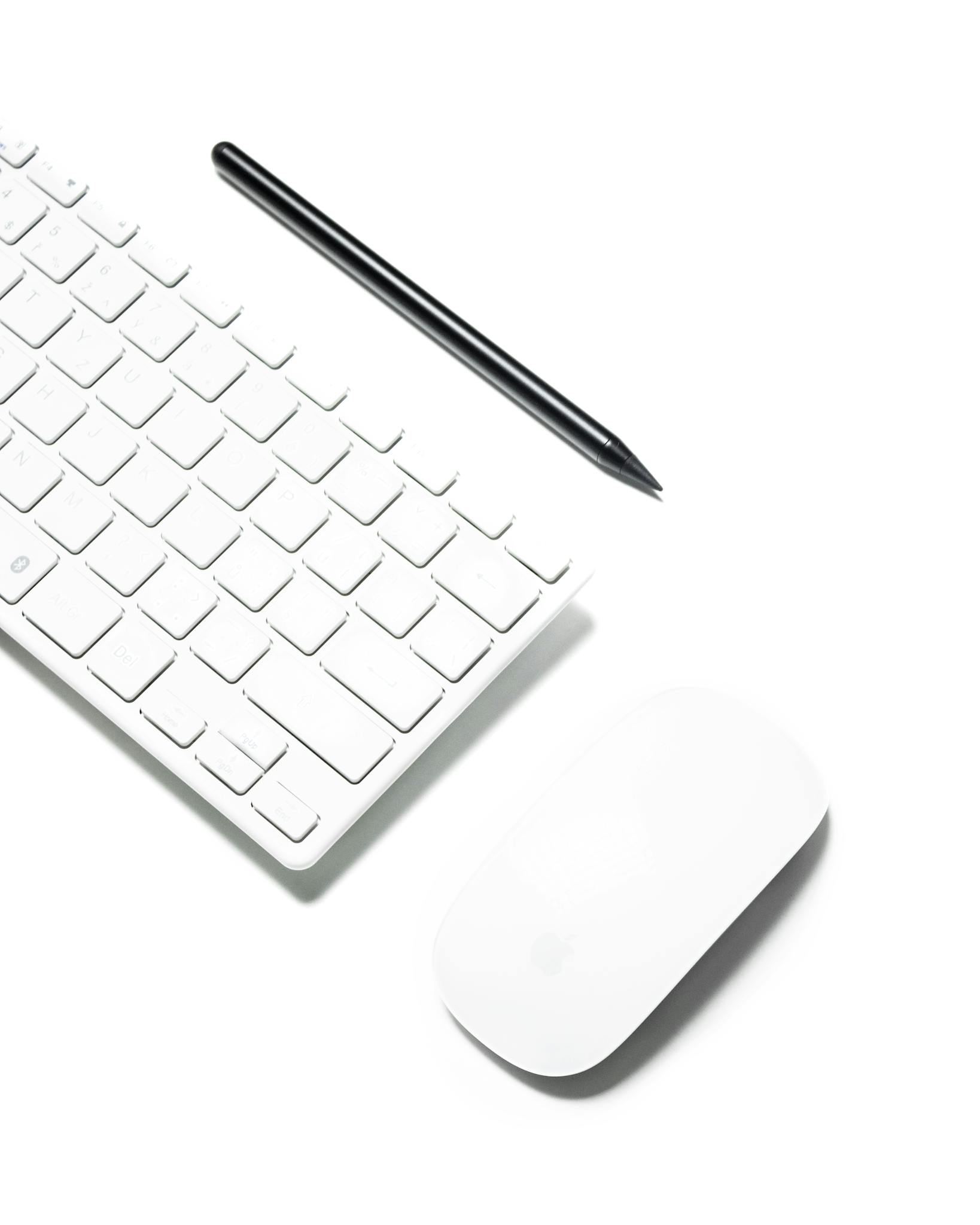 A clean and minimalist workspace featuring a wireless keyboard, mouse, and stylus on a white background.