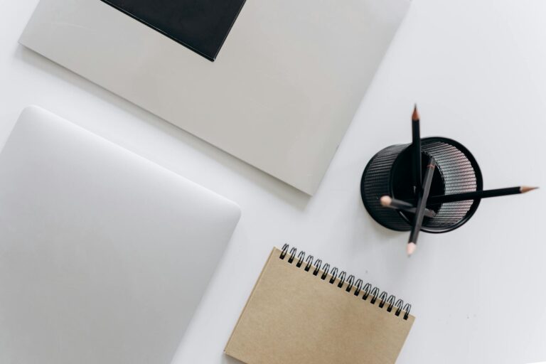 Top view of a minimalist workspace featuring a laptop, notepad, and stationery on a white surface.